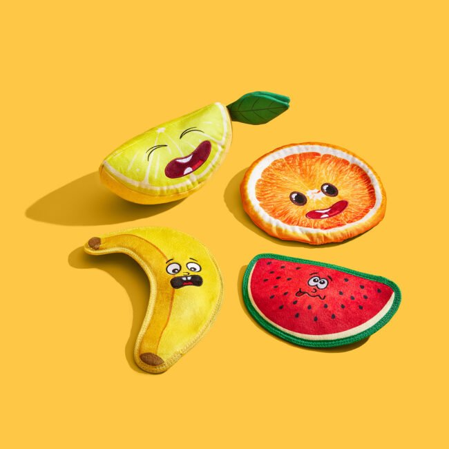 Studio product photography of fruit-shaped dog toys on a yellow background for a commercial pet brand campaign